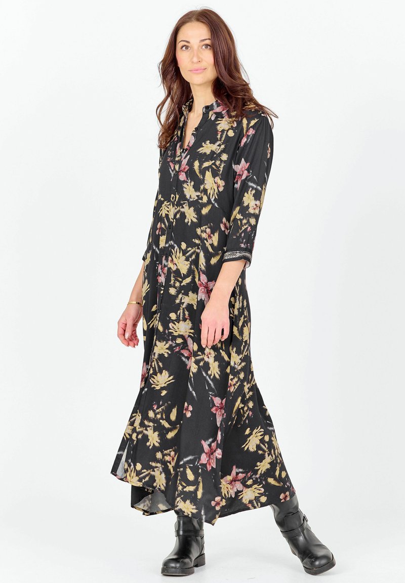 Femme portant une longue robe noire fleurie avec des manches trois-quarts et des bottines noires, debout devant un fond blanc.