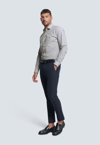 Herren Hemd mit Knopfleiste und geometrischem Muster in Weiß und Marineblau, kombiniert mit dunklen Hosen und schwarzen Mokassins, vor einem hellen Hintergrund stehend.