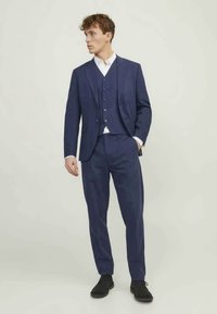 Fato azul-marinho de três peças, feito de um tecido texturizado, apresentando um blazer ajustado, colete e calças, combinado com uma camisa branca e sapatos pretos.