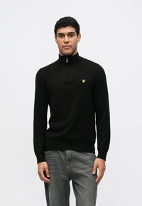 Fekete cipzáras nyakú pulóver bordázott gallérral és mandzsettákkal, a mellrészen egy kis sárga logóval. Szürke farmernadrággal viselve.