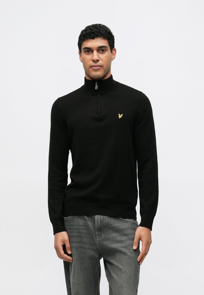 Fekete cipzáras nyakú pulóver bordázott gallérral és mandzsettákkal, a mellrészen egy kis sárga logóval. Szürke farmernadrággal viselve.