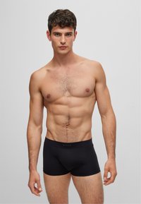 Bóxer negro con tejido suave y cinturilla elástica que presenta un logo. Modelo con una figura tonificada, posando directamente frente a la cámara.