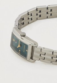 Ezüst fém karóra téglalap alakú tokkal, zöld számlappal, arany színű mutatókkal és fém szemes szíjjal. Állításhoz koronával rendelkezik.