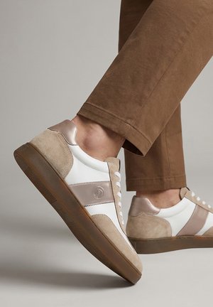 Person wearing beige and white low-top sneakers with brown rubber soles and brown pants, one foot lifted against a plain background.