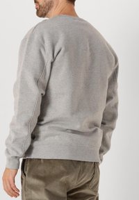 Sudadera gris con puños y dobladillo acanalados, con cuello redondo y costuras laterales. Se ve un par de pantalones de pana color oliva a la altura de la cintura.
