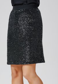 Jupe en sequins noirs avec une taille ajustée, dotée de plis verticaux et d'une coupe au genou, mise en valeur par des sequins réfléchissants sur toute sa surface.