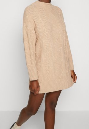 Robe-pull beige en maille torsadée à manches longues, portée avec des chaussettes beiges et des chaussures noires sur un fond clair uni.