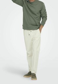Felpa verde oliva con collo a giro, taglio rilassato; abbinata a pantaloni beige chiaro con coulisse e scarpe verde scuro con suola beige.
