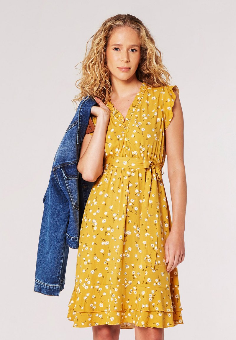 Gelbes Blumen Kleid mit kurzen Rüschensäumen, einem V-Ausschnitt und einem Taillengürtel, kombiniert mit einer blauen Jeansjacke.