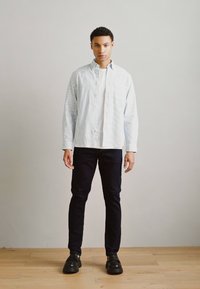 Camisa de botón a rayas azul claro, camiseta blanca debajo, jeans oscuros y zapatos negros, de pie sobre un suelo de madera contra un fondo liso.