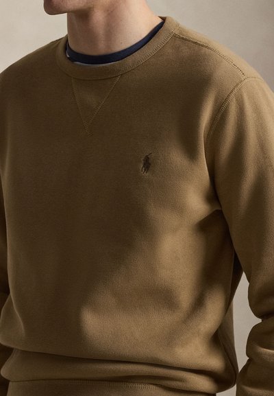 Άνδρας που φοράει καφέ φούτερ με στρογγυλή λαιμόκοψη και μικρό κεντητό λογότυπο στο στήθος, από πάνω από μπλε σκούρο πουκάμισο.