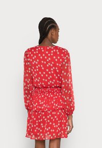 Vestido floral vermelho com mangas longas, apresentando uma saia em camadas, cintura elástica e margaridas brancas numa tela transparente. Vista traseira apresentada.