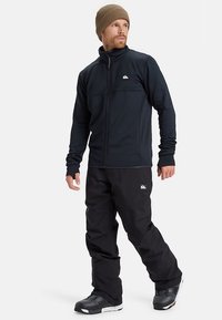 Μαύρο φερμουάρ μπουφάν με ψηλό γιακά, συνδυασμένο με μαύρα παντελόνια. Ο άνδρας φοράει ένα καφέ σκουφάκι και χιονομπότες, με ένα λογότυπο και στα δύο ρούχα.