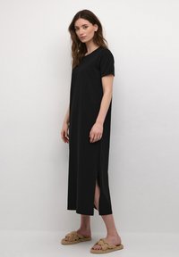Black, knee-length t-shirt dress with short sleeves, side slits, smooth texture, and a relaxed fit. Worn with knotted slide sandals.
