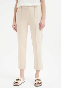 Beige op maat gemaakte broeken met een rechte snit, voorzien van voorplooien en een gladde textuur. Draagt met witte instap sandalen met gouden accenten.