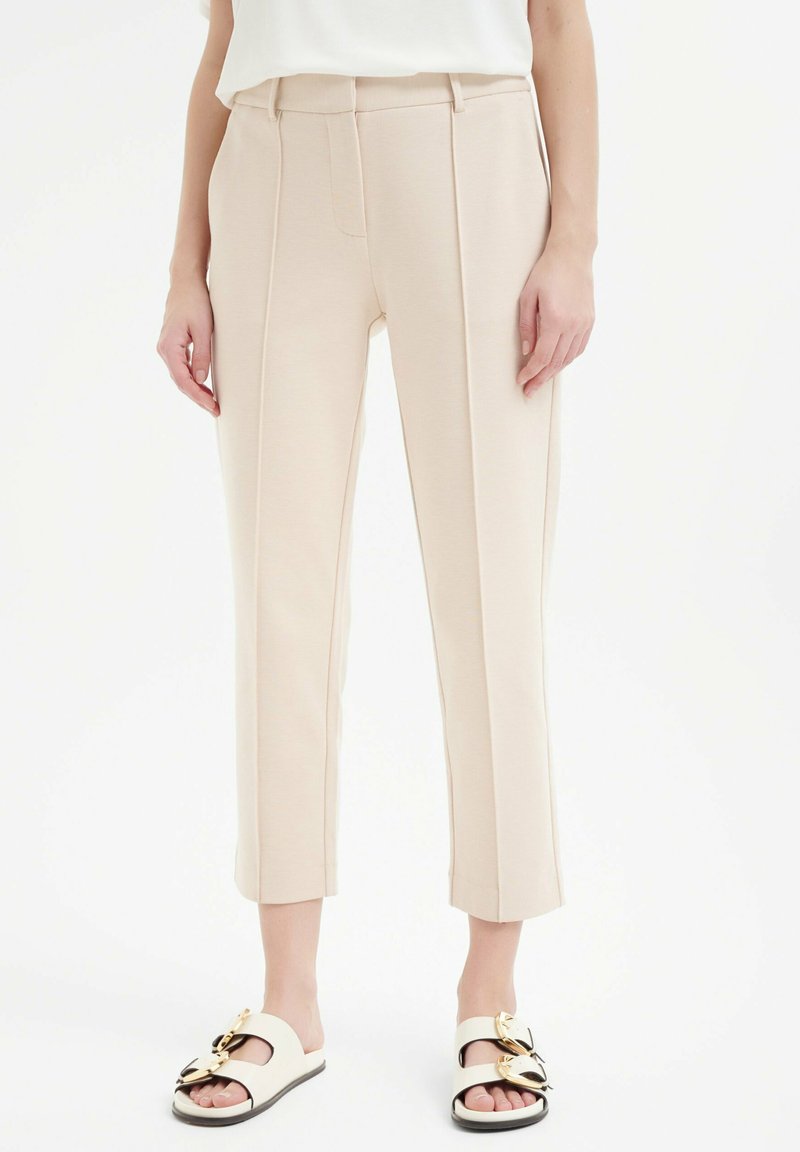 Beige op maat gemaakte broeken met een rechte snit, voorzien van voorplooien en een gladde textuur. Draagt met witte instap sandalen met gouden accenten.
