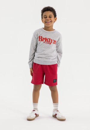 Grijze sweatshirt met rode "Brklyn" tekst, gecombineerd met rode shorts. Kind draagt witte sneakers met bruine zolen en grijs-wit gestreepte sokken.