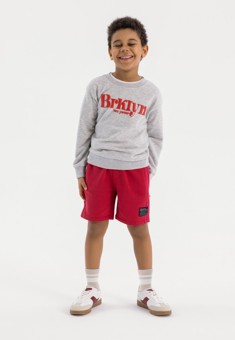 Sweat-shirt gris avec le texte "Brklyn" en rouge, associé à un short rouge. Enfant portant des baskets blanches avec des semelles marron et des chaussettes rayées gris-blanc.