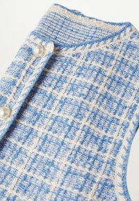 Niebiesko-biała kamizelka o wzorzystym dzianym materiale z fakturowaną powierzchnią, ozdobiona perłowymi guzikami i kołnierzykiem. Widoczne detale przeszyć.