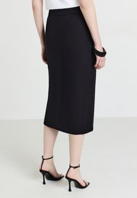 Schwarzer Midirock aus glattem Material, mit tailliertem Bund und geradem Schnitt. Kombiniert mit schwarzen High-Heelsandalen mit Knöchelriemen.