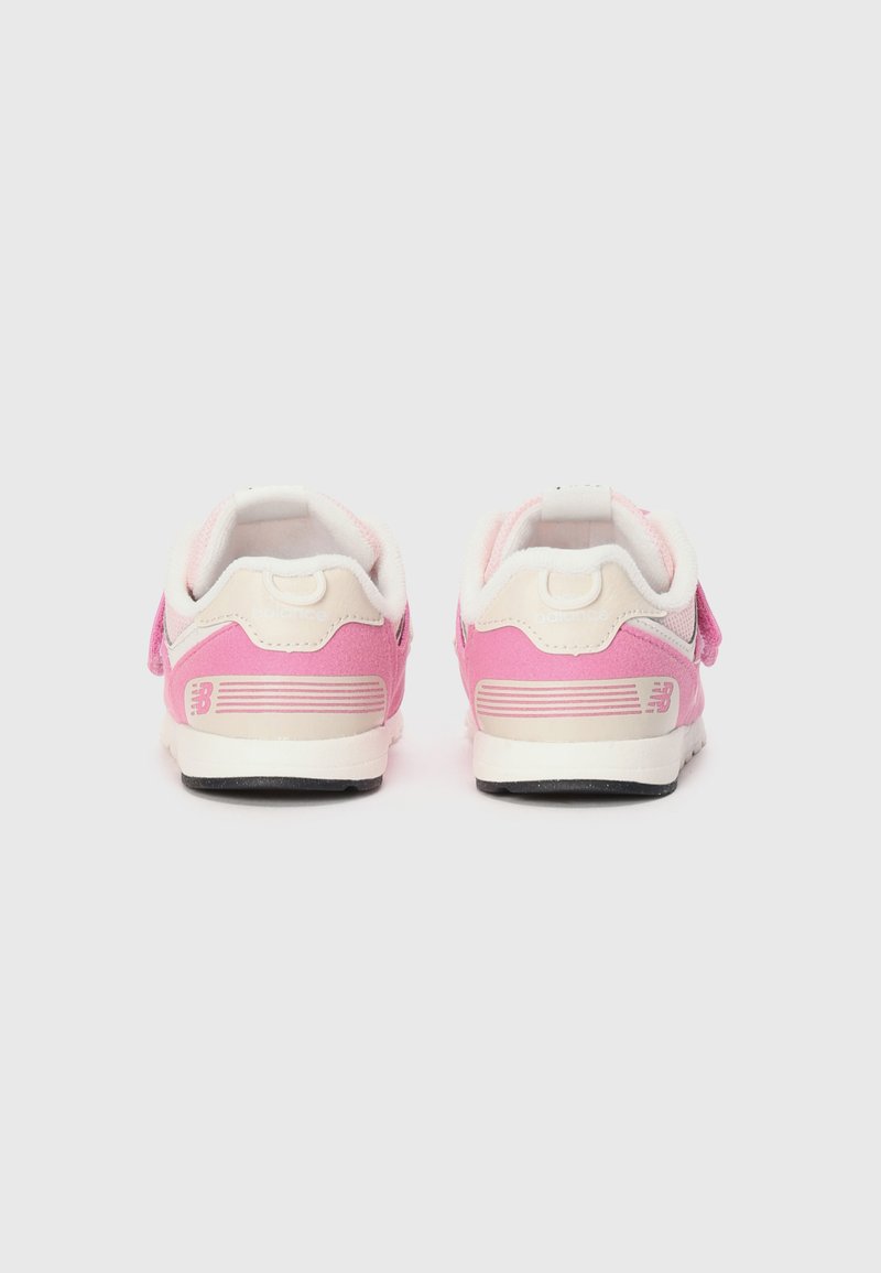Un par de zapatillas pequeñas en rosa y blanco de New Balance vistas desde atrás sobre un fondo gris claro liso.