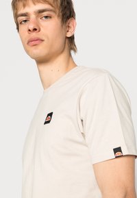 Light beige t-shirt made of cotton, featuring a small black logo patch with a colorful design on the left chest and sleeve.