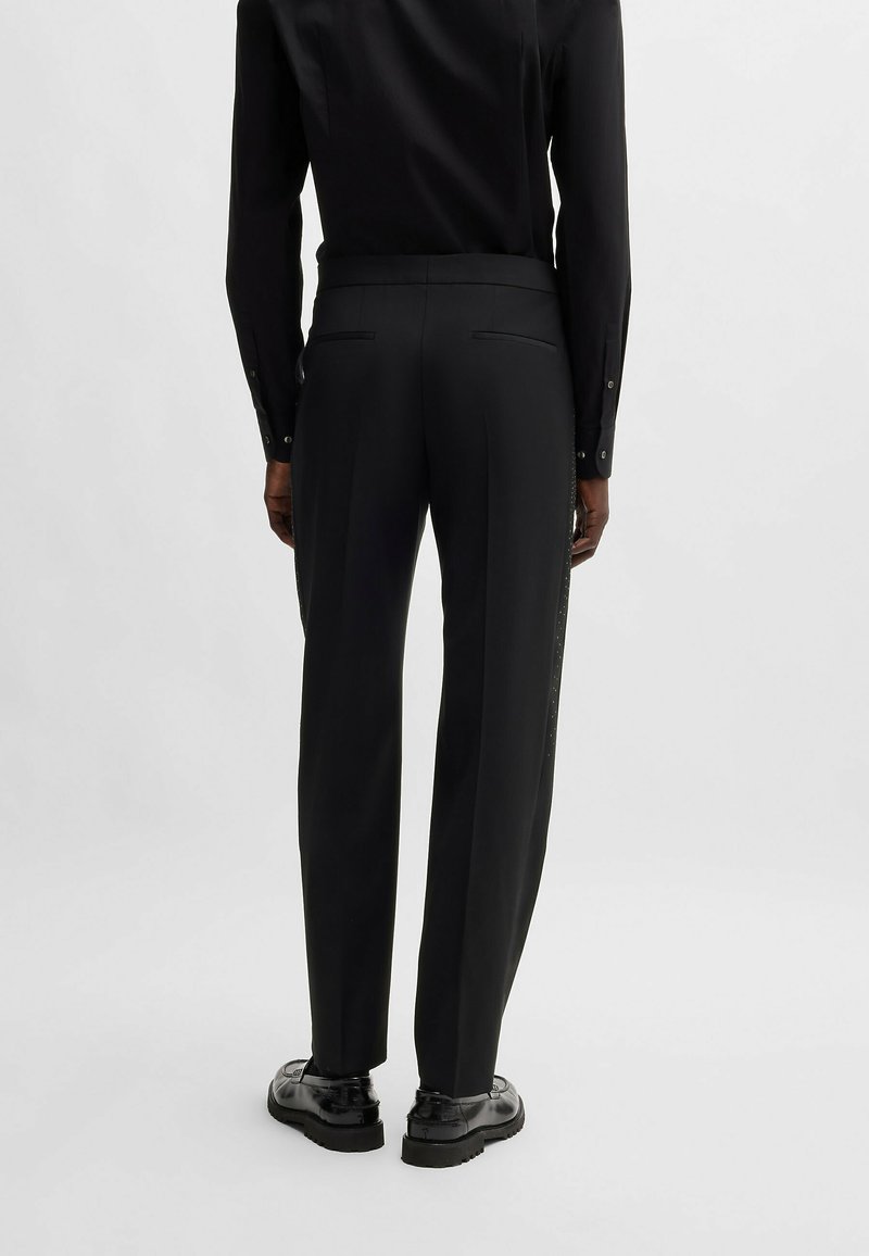 Person, der schwarze maßgeschneiderte Hosen, ein schwarzes Hemd und schwarze polierte Schuhe trägt, steht vor einem schlichten hellen Hintergrund.