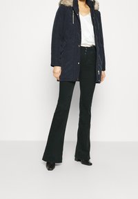 Navy parka with faux fur hood, zipper and snap closures, features side pockets and is paired with dark flared pants and black heels.