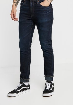 Mand iført mørke slim-fit jeans med opkørte manchetter og sorte lave sneakers med hvide snørebånd og sål, står med den ene hånd i lommen.