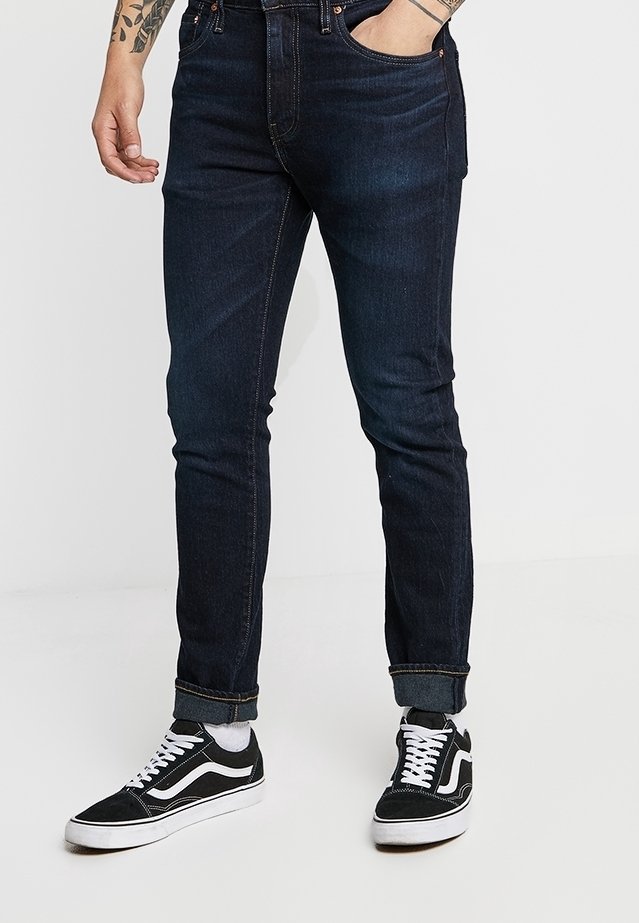 Homme portant un jean slim foncé avec les revers remontés et des baskets basses noires avec des lacets blancs et une semelle blanche, debout avec une main dans la poche.
