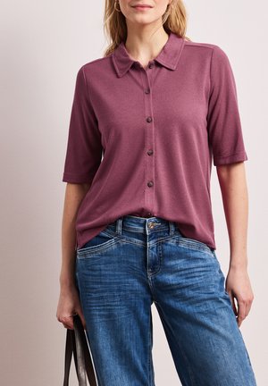 Kobieta w mauve koszuli na guziki z krótkim rękawem, włożonej do niebieskich dżinsów, trzymająca czarną torebkę przy boku.