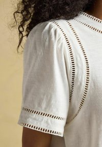 Valge lühikeste varrukatega särk, mis on valmistatud kergetest materjalidest ja millel on dekoratiivsed lõikeväljad õmbluste ja varrukate ääres. Sujuv tekstuur.