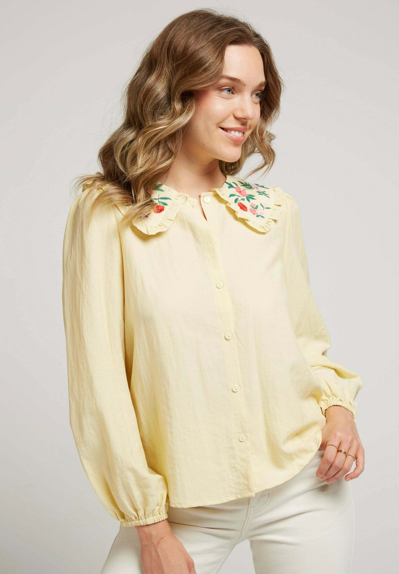 Blouse jaune à boutons avec un col volanté orné de broderies florales. Manches longues avec poignets élastiques, fabriquée dans un tissu léger.