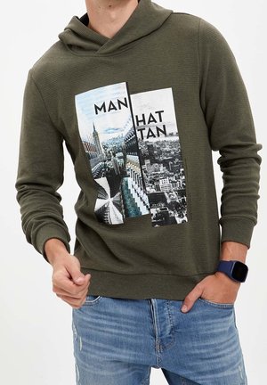 Mężczyzna w oliwkowo-zielonej bluzie z kapturem z grafiką miasta Nowy Jork i słowem "Manhattan", w połączeniu z jasnoniebieskimi dżinsami i ciemnym zegarkiem na nadgarstek.