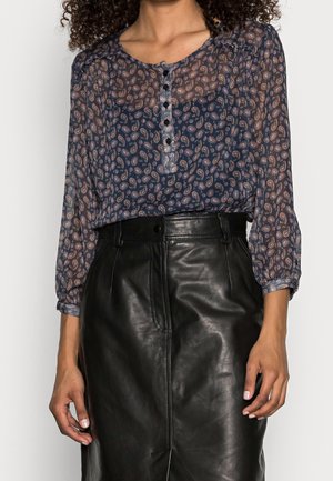 Femme portant un chemisier transparent bleu marine à motif cachemire avec des boutons noirs et une jupe en cuir noire taille haute avec des détails de coutures à l'avant.