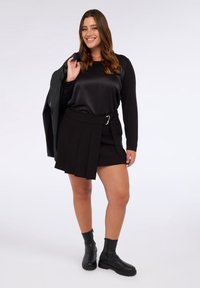 Schwarzes Satin-Langarmoberteil kombiniert mit einem schwarzen plissierten Rock mit Gürtel. Das Model trägt schwarze Stiefeletten und hält eine grüne Jacke.