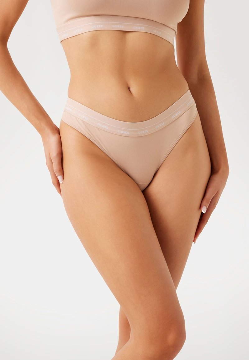 Beige tanga alsónemű sima textúrával és diszkrét elasztikus derékszallaggal, amelyen logó található. A kivágás minimalista, a formához illeszkedő dizájn hangsúlyozásával.