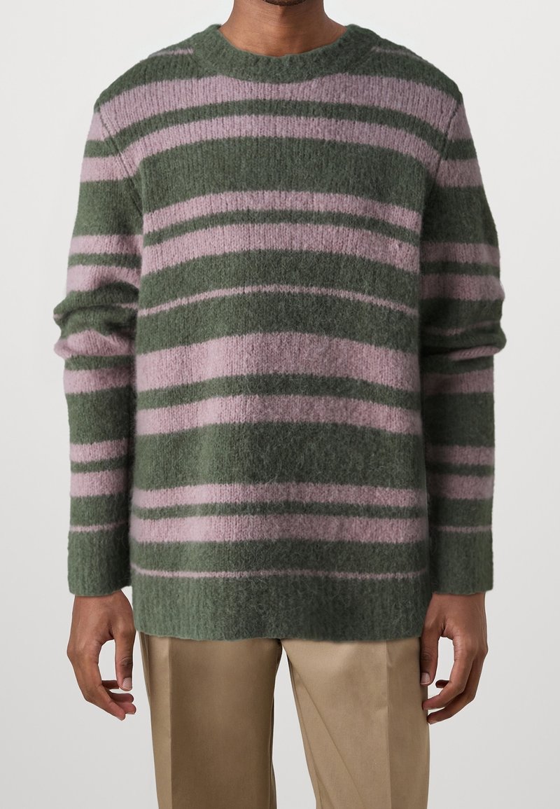 Homme portant un pull duveteux à rayures horizontales vertes et violettes avec un pantalon beige, debout devant un fond uni.