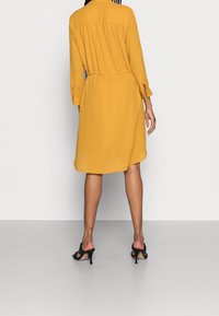 Vestido camisero amarillo con pequeños lunares blancos, corte relajado, cinturilla elástica, mangas largas y dobladillo curvado. Llevado con sandalias negras de tacón.
