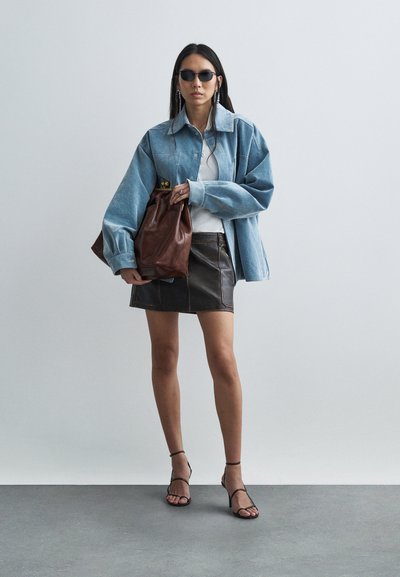 Žena v nadměrné modré džínové bundě, bílém topu, černé kožené sukni, černých sandálech na podpatku s pásky, tmavých slunečních brýlích, držící velkou hnědou koženou tašku.