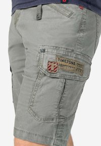 Gray cargo shorts with multiple pockets, featuring a logo patch and decorative stitching. Material appears lightweight and durable.