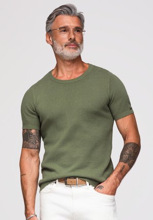 Homme aux cheveux gris et à la barbe portant des lunettes transparentes, une chemise verte texturée à manches courtes, un pantalon blanc et des tatouages sur les deux bras, posant avec la main dans la poche.