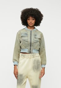 Skrajšani denim bomberska jakna v svetlo modri barvi z zadrgo, dvema žepoma na prsih in rebrastimi manšetami. V kombinaciji s bež, poškrabljenimi trenirkami.