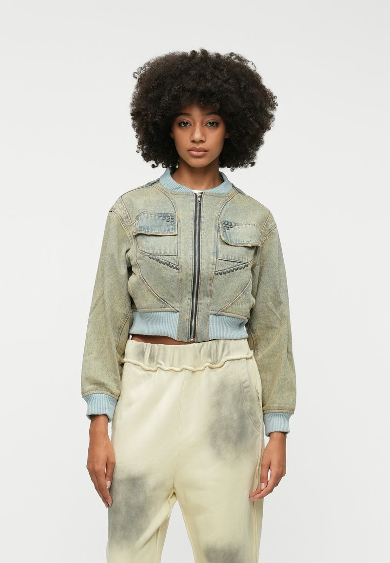 Skrajšani denim bomberska jakna v svetlo modri barvi z zadrgo, dvema žepoma na prsih in rebrastimi manšetami. V kombinaciji s bež, poškrabljenimi trenirkami.