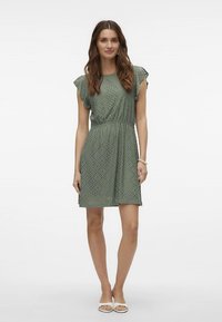 Zielona sukienka z ażurowymi detalami, krótkie falbaniaste rękawy, elastyczna talia i luźny krój, sięgająca ponad kolana. Noszona z białymi sandałami.