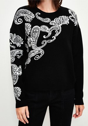 Sort sweater med hvide blomster- og paisleymønstre på skulder, bryst og ærme, båret sammen med sorte bukser.
