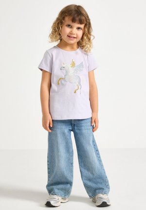 Ung pige med krøllet hår iført en lys lilla skjorte med en paliet Pegasus og løse blå jeans, står og smiler mod en ensfarvet baggrund.