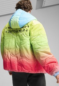 Töltött pufferdzseki, zöldtől rózsaszínig terjedő gradient mintával, kék kapucnival és hímzett szöveggel a hátsó részén.