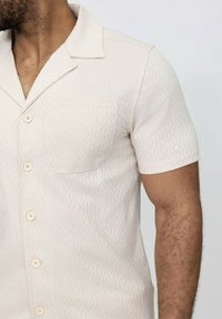Chemise à manches courtes, de couleur beige clair, présentant un motif zébré subtil, un col classique et une poche plaquée ; ornée de boutons clairs.