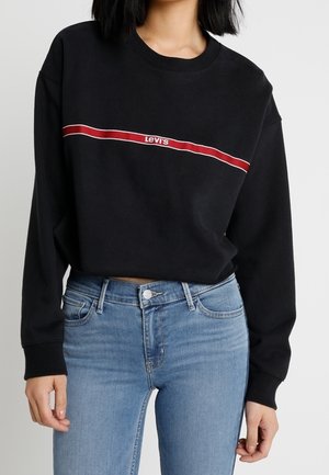 Žena, která má na sobě černou krátkou mikinu Levi's s červeným a bílým pruhem přes hrudník a světle modré džíny s vysokým pasem.